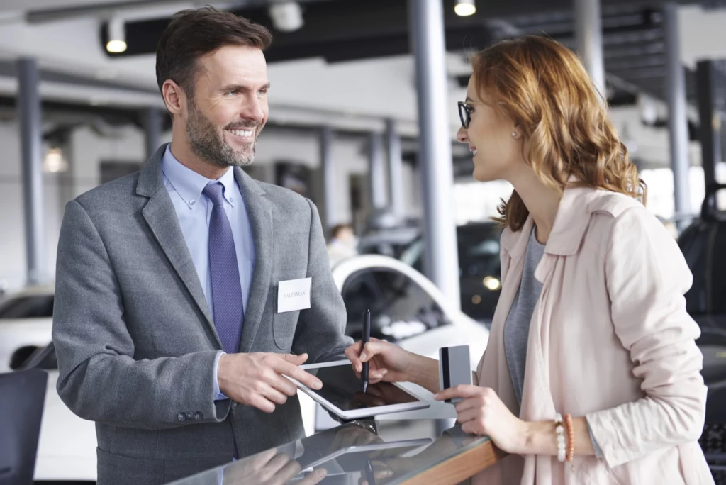 Homme blanc en train de faire signer un document sur une tablette électronique à une femme blanche et rousse, dans un lieu de vente de voiture.
